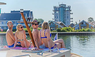 Zwei Männer und Frauen sitzen auf einem Boot. Im Hintergrund sieht man den Müritzturm im Hafendorf Müritz. 