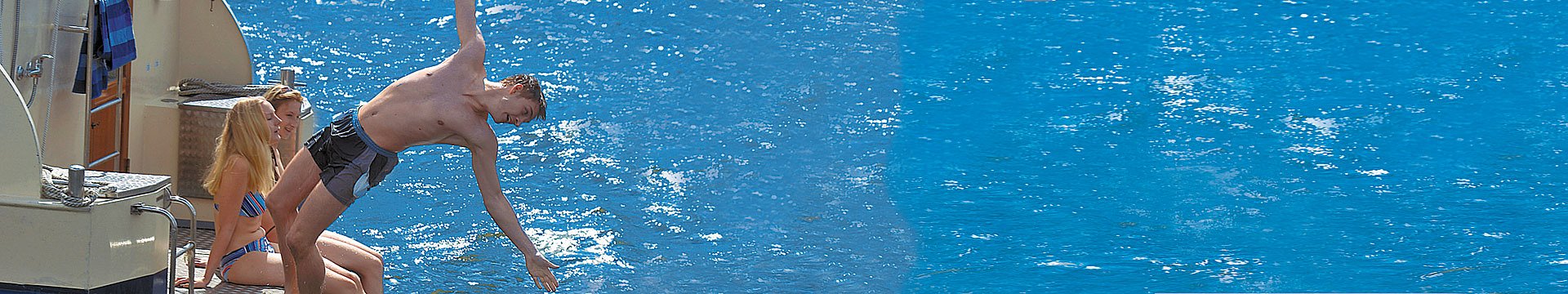 Drei Jugendliche sitzen auf einer Badeplattform, ein junger Mann springt lustig ins Wasser.