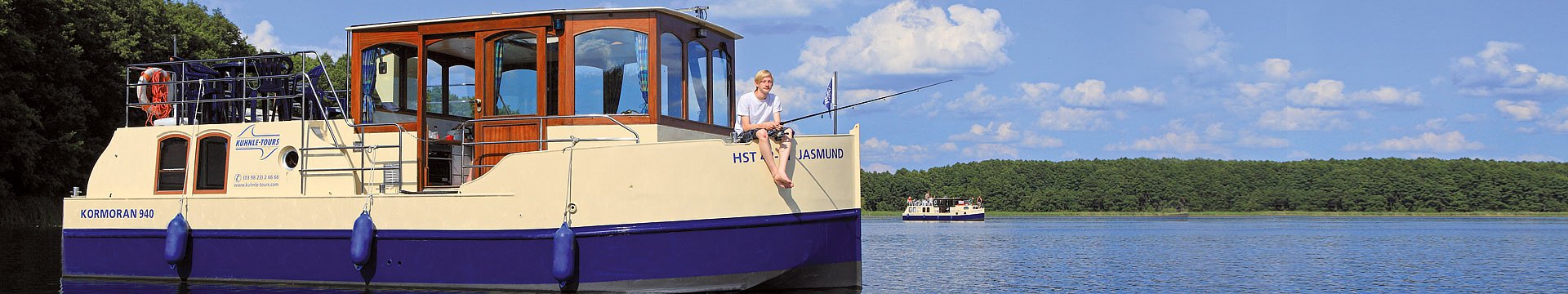 Ein Hausboot, Marke Kormoran, ankert auf einem See in der Nähe des Ufers. Ein junger Mann sitzt gemütlich an Deck und angelt.