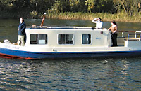 Ein Hausboot im Stil eines englischen narrow-boats fährt über einen Waldsee.