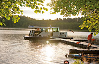 Ein Hausboot hält an einem Steg in romantischer Atmosphäre. Die Sonne spiegelt sich im See.