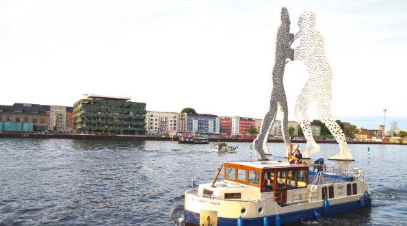 Das Hausboot Kormoran fährt in Berlin am Kunstwerk molecule man vorbei